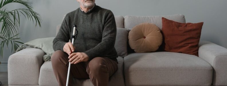 Man sitting on a couch wearing sun glasses, holding a white walking cane